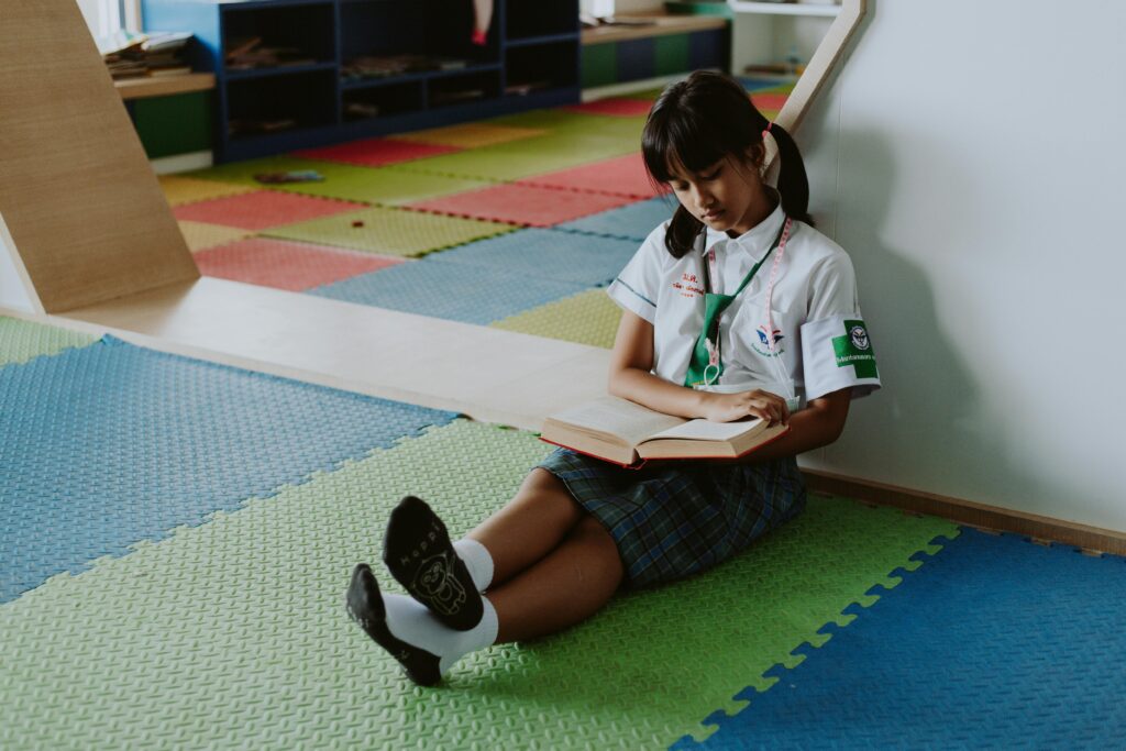 pexels-photo-10643451-10643451 A young girl in school uniform sits on colorful mats reading a book indoors.
