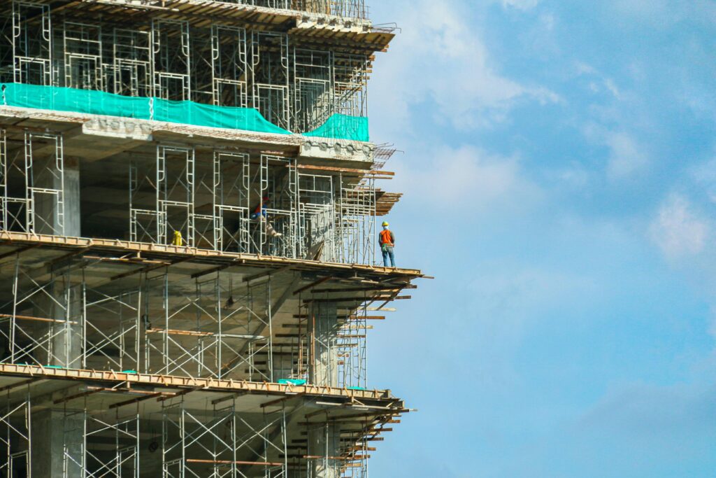 pexels-photo-4966809-4966809 A construction worker stands on a scaffolding at a high-rise building under a blue sky.